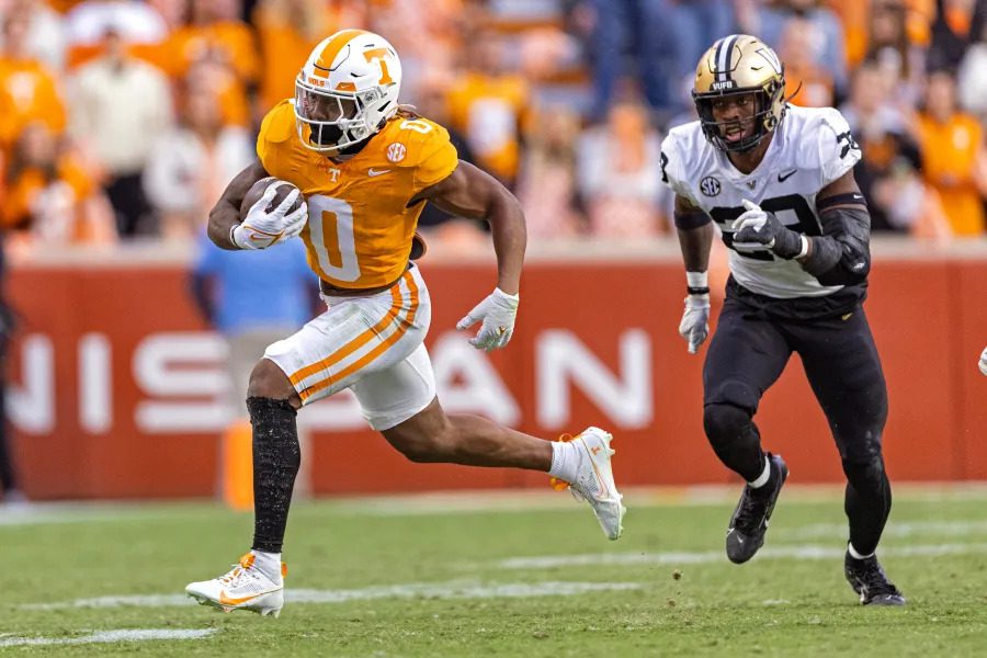 Trendsnip A Tennessee Volunteers football player in an orange jersey runs with the ball, pursued by a Vanderbilt Commodores player in a black and white jersey during a high-stakes game.